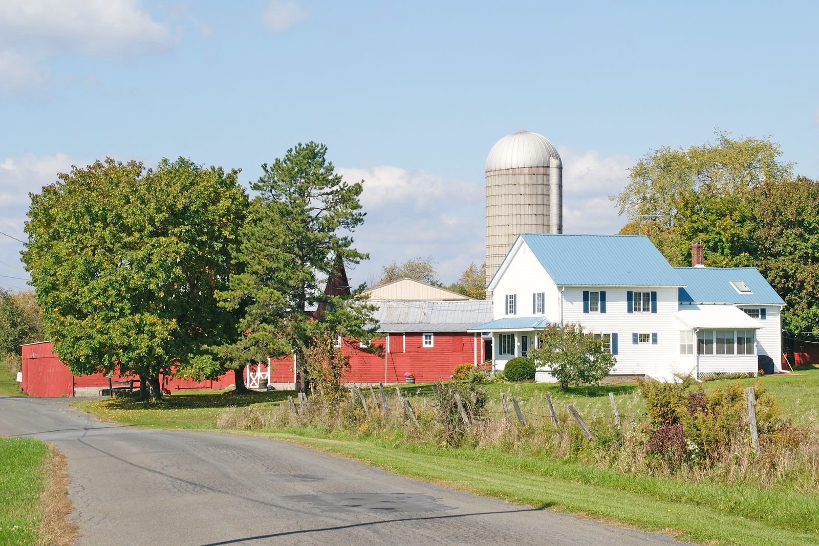 A two-lane blacktop road curves past a white farmhouse, red barns, and a silo in upstate New York.