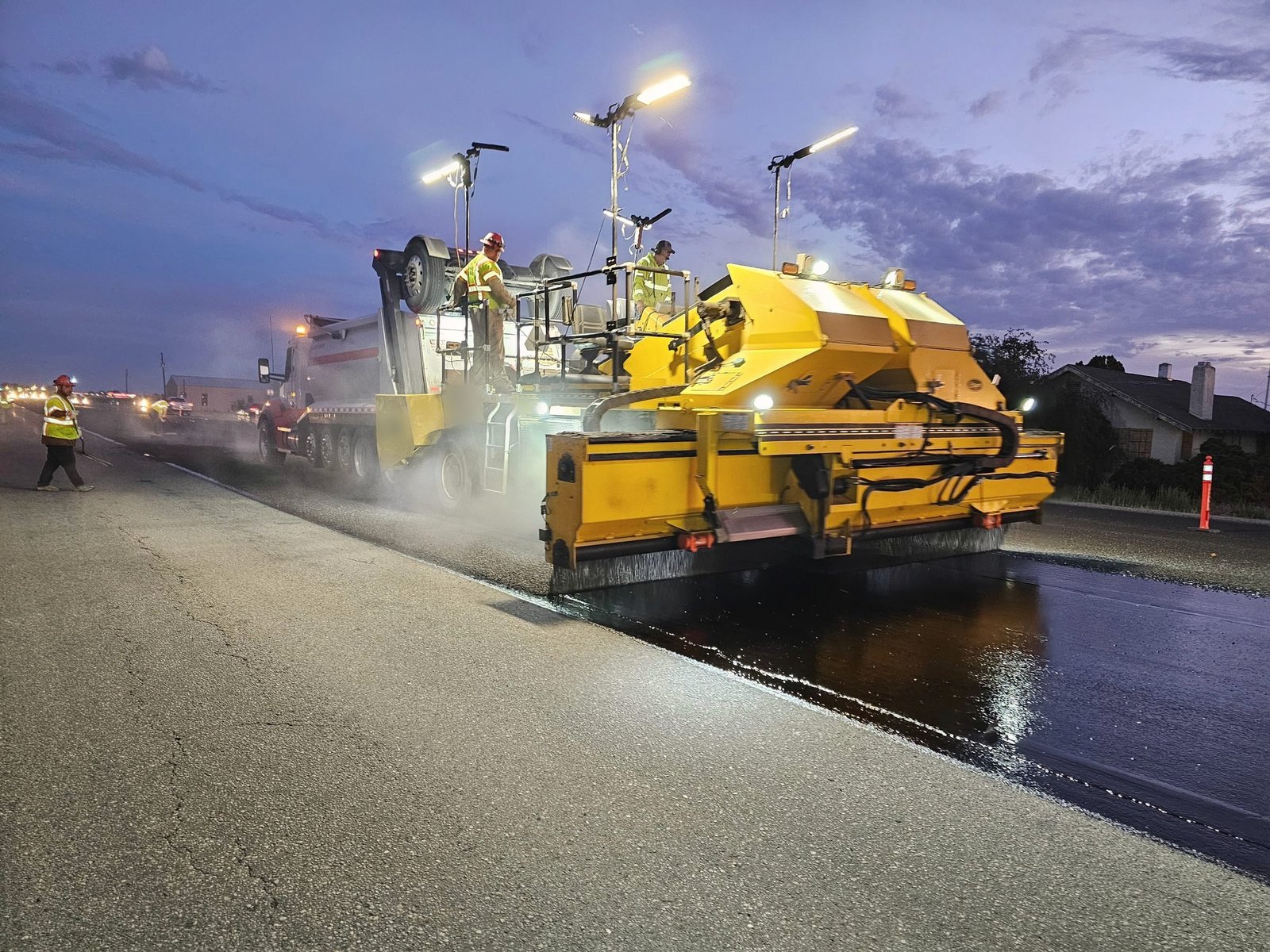A paving crew lays hot asphalt at dusk, work lights on and steam rising off the fresh mat.