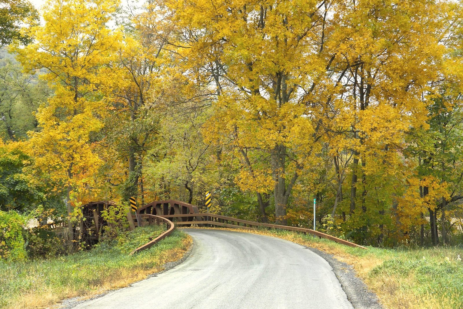 A two-lane blacktop road curves past an iron bridge under yellow fall trees near Feura Bush, New York.